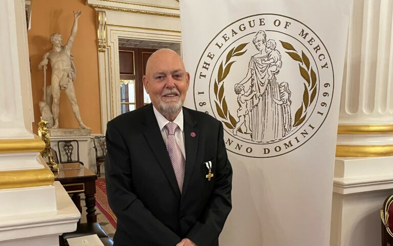 Ray Baglee wearing his award, standing in front of a League of Mercy banner
