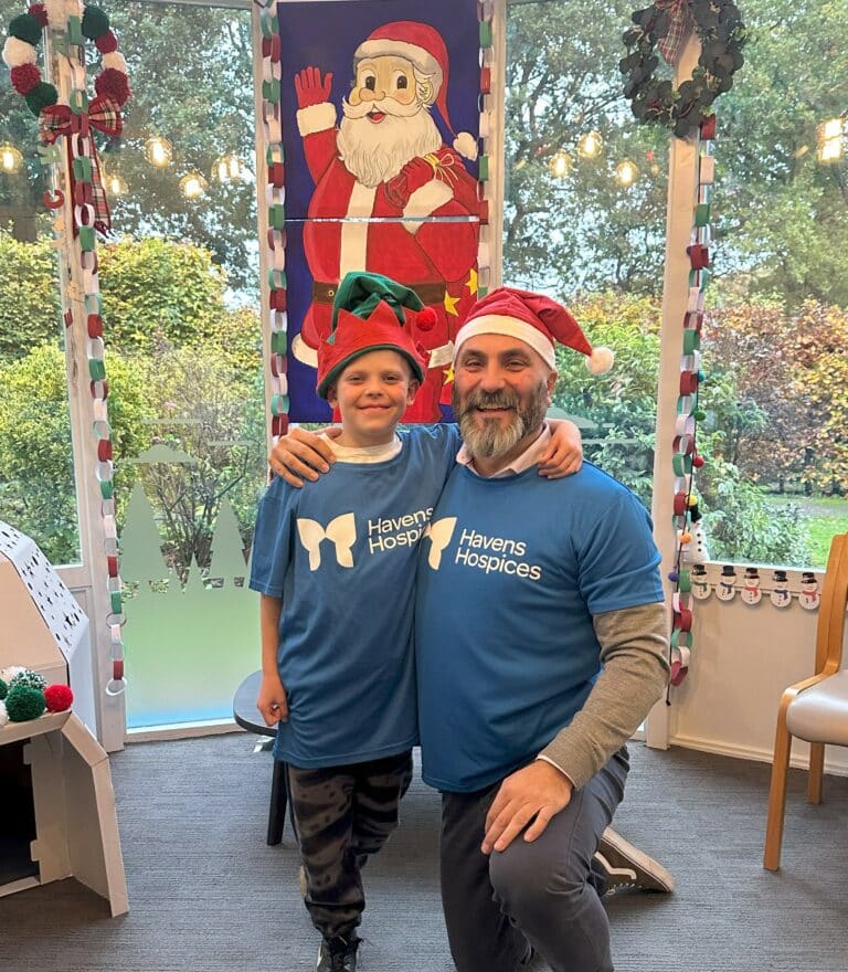 Jack and Rob wearing Havens Hospices t-shirts and elf and Santa hats in front of a festive backdrop