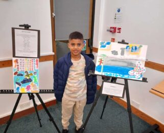 A young boy standing next to his paintings smiling