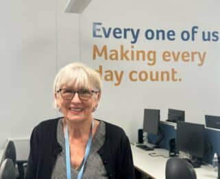 Volunteer, Diane, in Havens Hospices office, smiling in front of the "Every one of us Making every day count" sign.