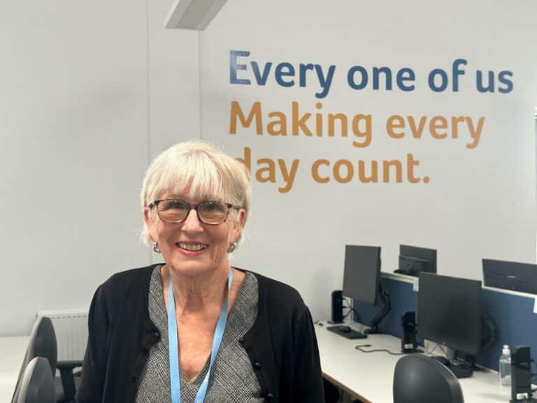 Volunteer, Diane, in Havens Hospices office, smiling in front of the "Every one of us Making every day count" sign.