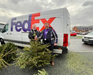 Two male FedEx workers smiling, holding a Christmas tree in front of a FedEx van