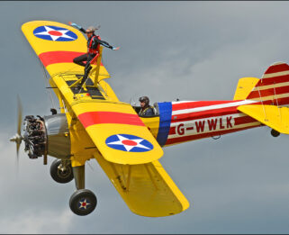 Woman standing on top of a flying aeroplane, her arms are outstretched.