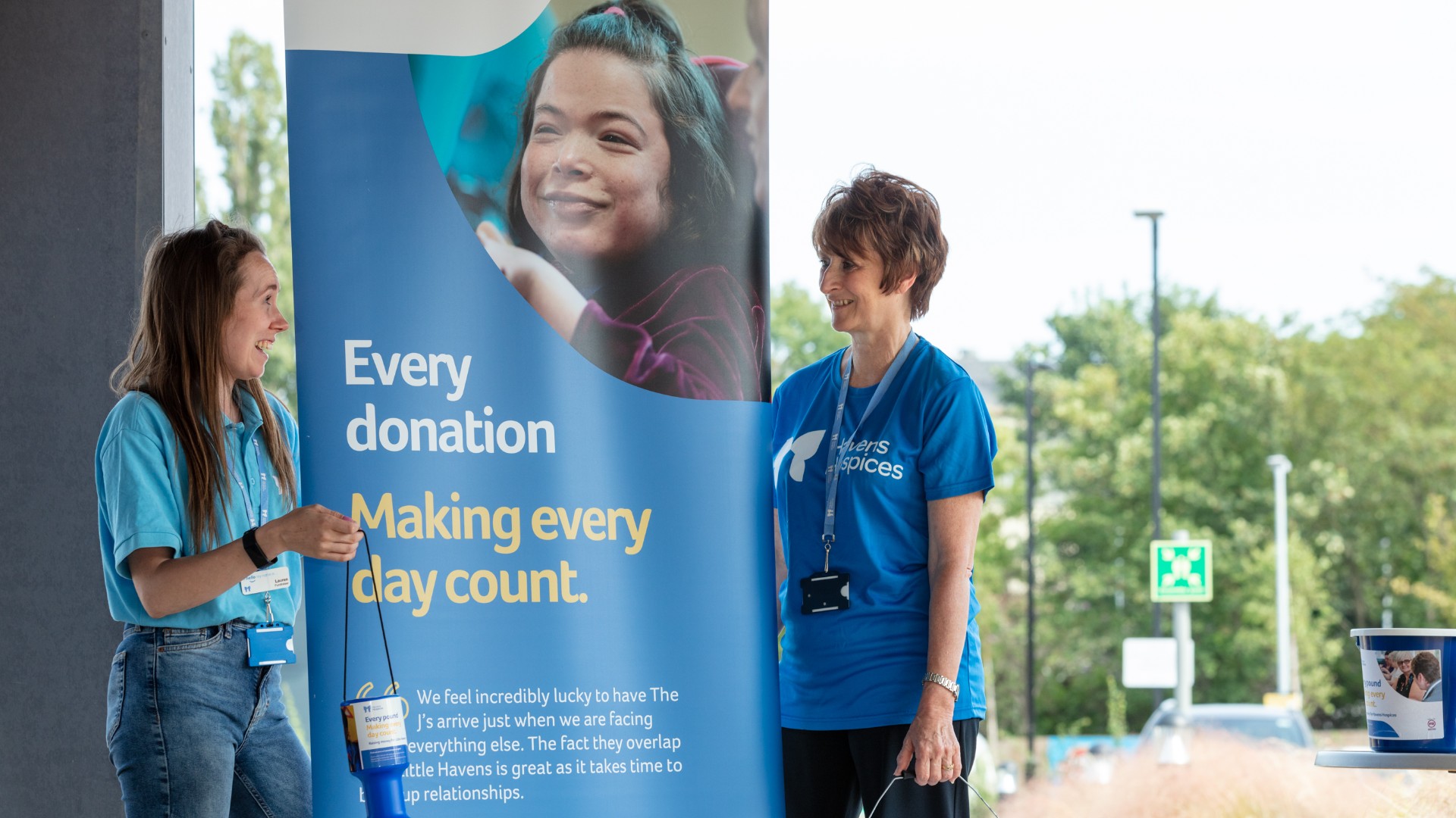 Two Havens Hospices Fundraisers in blue branded 'Havens Hospices' t-shirts stand either side of a banner reading 'Every donation, Making every day count'
