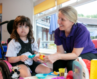 A young child sat in her wheelchair with pink wheels looking direct at camera while next to her is a HCA wearing a purple top looking at the child. In front of them is playdough on the table