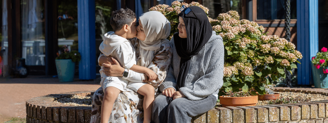 Zak's mum is wearing a cream headscarf kissing Zak's face, sat next to them outside is a teenage girl looking at the pair also wearing a black headscarf and glasses on her head