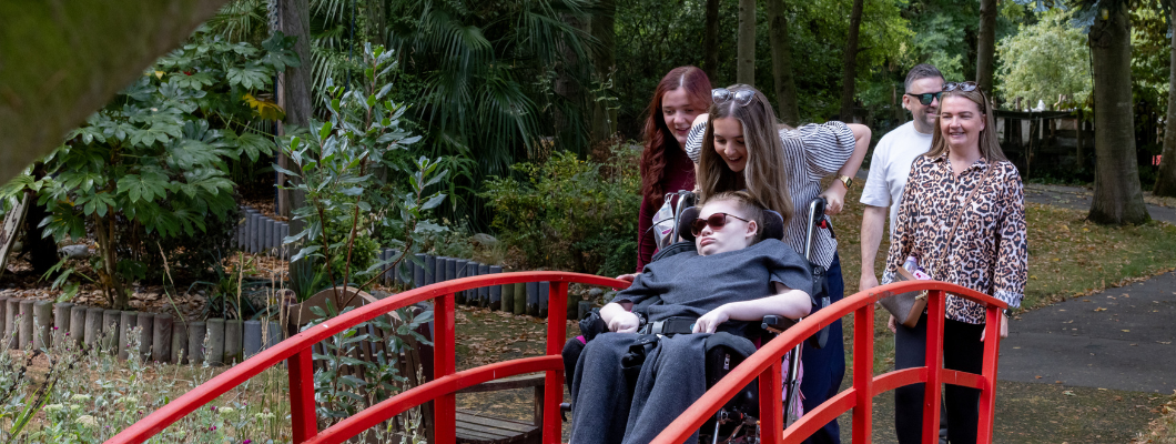 Teenage girl wearing sunglasses in a wheelchair being pushed over a red bridge in the garden