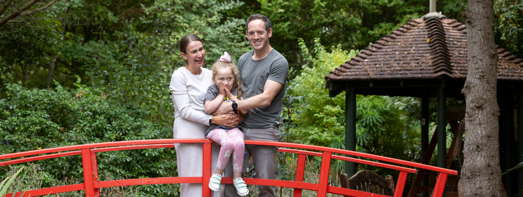 A family of three stands on a red wooden bridge in a garden, with both parents holding their young daughter between them.