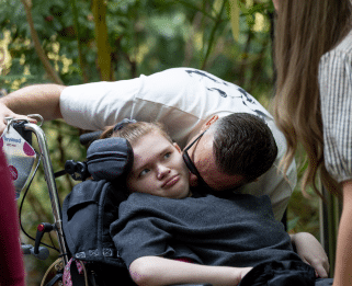 Dad leaning in to kiss face of teenage girl who is in a wheelchair and has her head turned to the camera