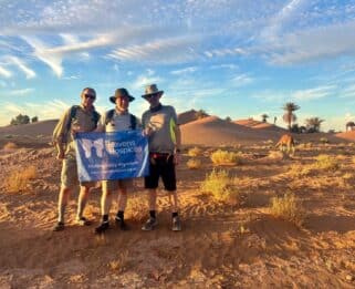Three supporters of Havens Hopsices standing in the Sahara Desert holding a Havens Hospices flag.