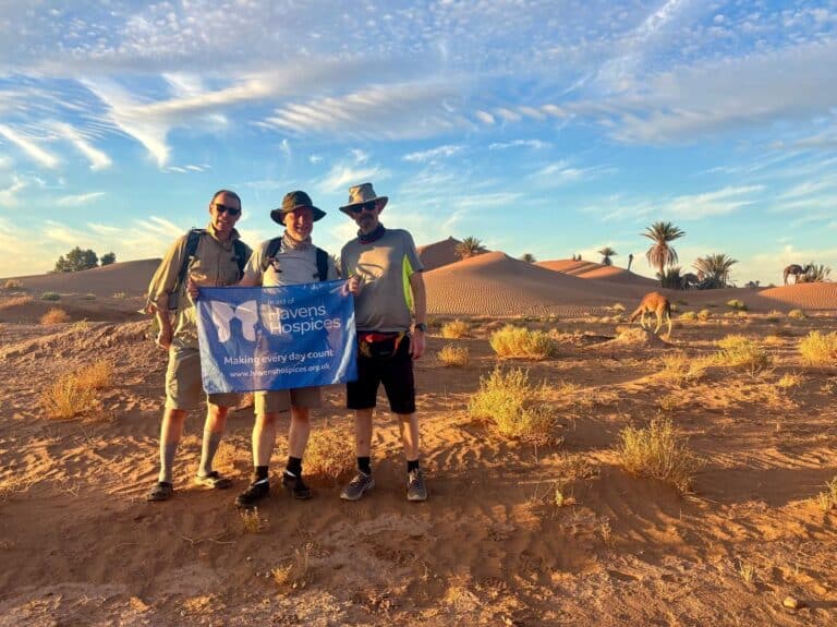 Three supporters of Havens Hopsices standing in the Sahara Desert holding a Havens Hospices flag.