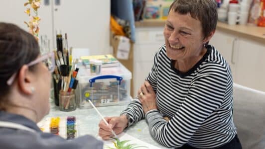 Lady in a striped top sat painting and smiling looking at a member of staff in a grey uniform