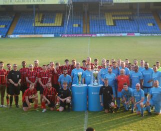 Two teams of men standing on the pitch of Root Hall with two trophies in the foreground
