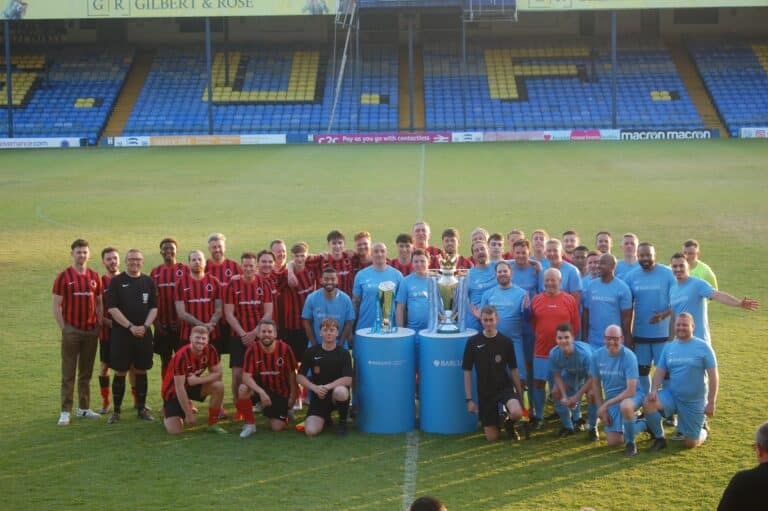 Two teams of men standing on the pitch of Root Hall with two trophies in the foreground