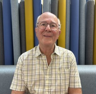 Man wearing a yellow checked shirt sat with coloured wall behind him