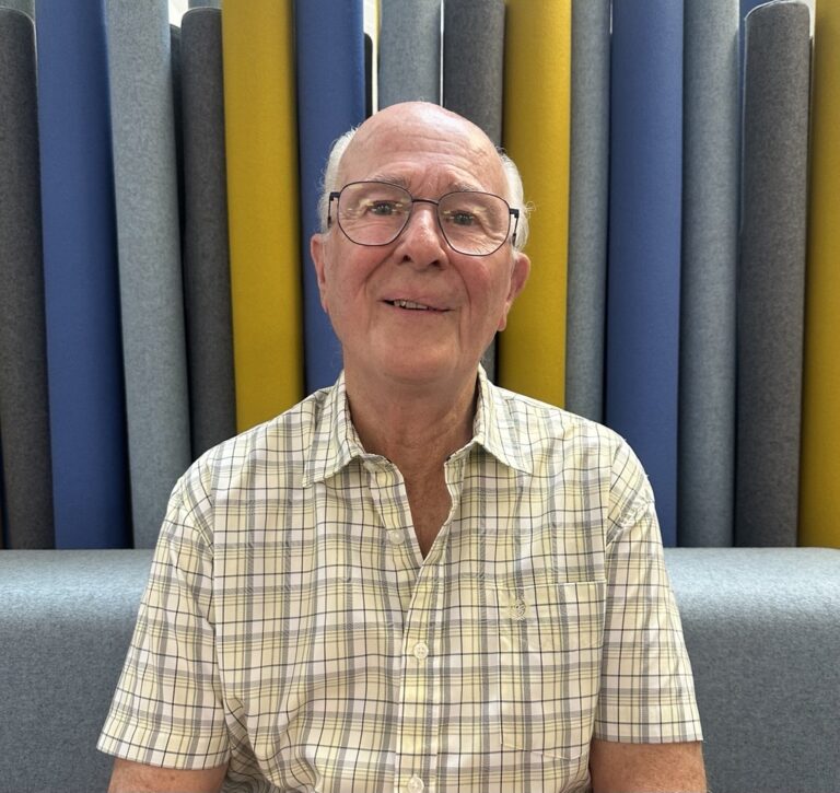 Man wearing a yellow checked shirt sat with coloured wall behind him