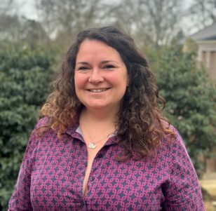 Lady wearing a purple patterns dress with curly brown hair down past her shoulders smiling at camera
