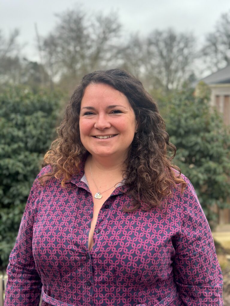 Lady wearing a purple patterns dress with curly brown hair down past her shoulders smiling at camera