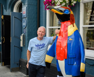 A man volunteering for the Waddle-on-Sea art trail leaning on a painted penguin sculpture.