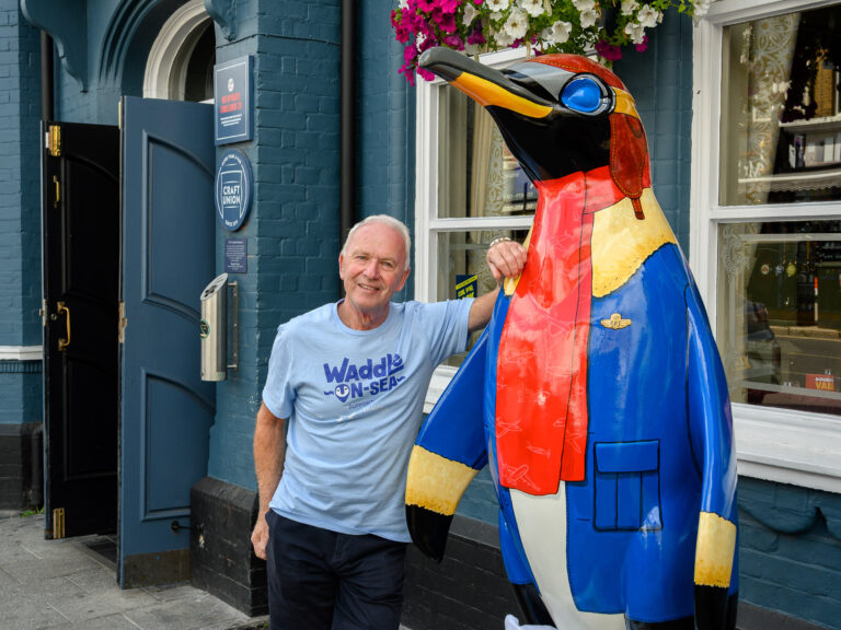 A man volunteering for the Waddle-on-Sea art trail leaning on a painted penguin sculpture.