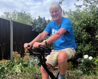 Nick Hart wearing a branded Havens Hospices t-shirt, sitting on his exercise bike in his garden