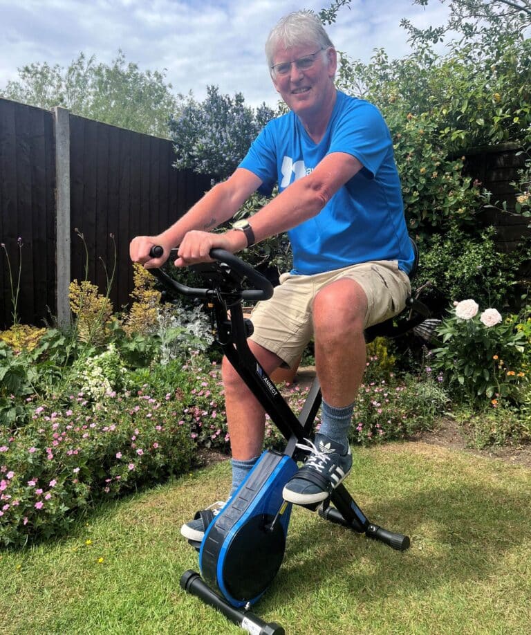 Nick Hart wearing a branded Havens Hospices t-shirt, sitting on his exercise bike in his garden