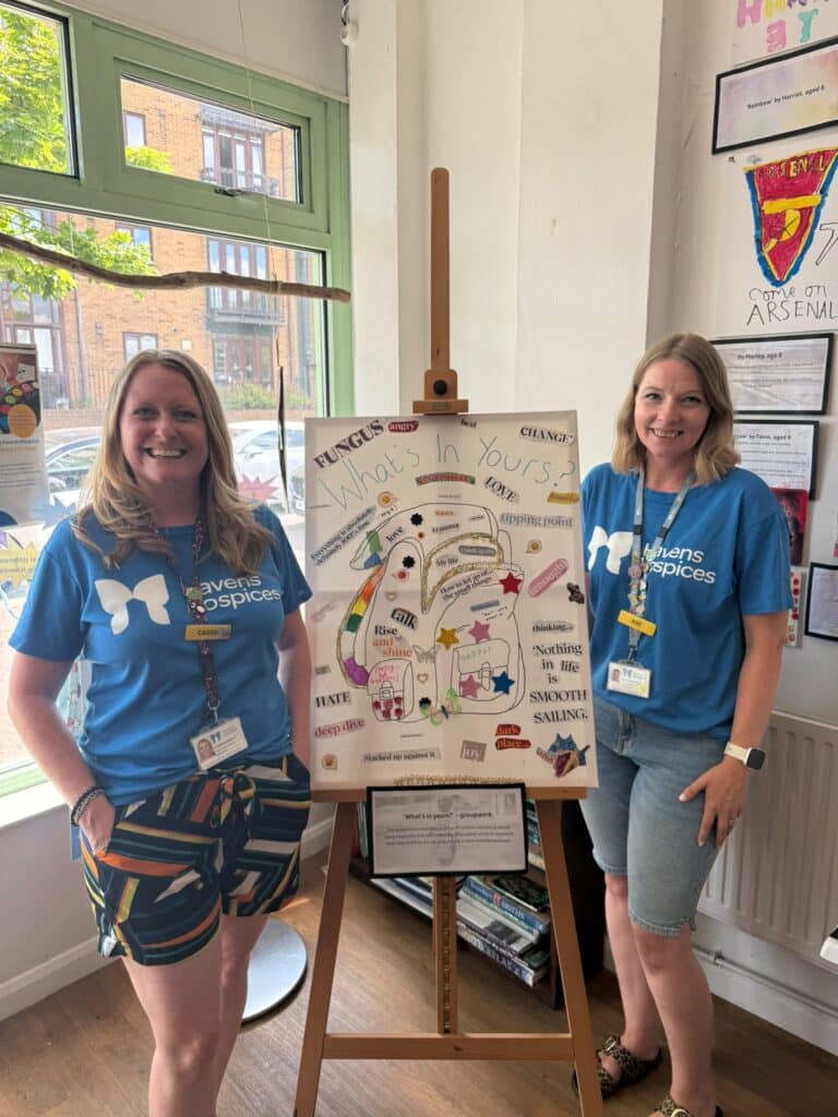 Two women wearing Havens Hospices tops standing next to an easel with a canvas on it.