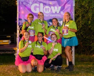 seven women smiling in bright clothing in front of a Glow for Havens banner