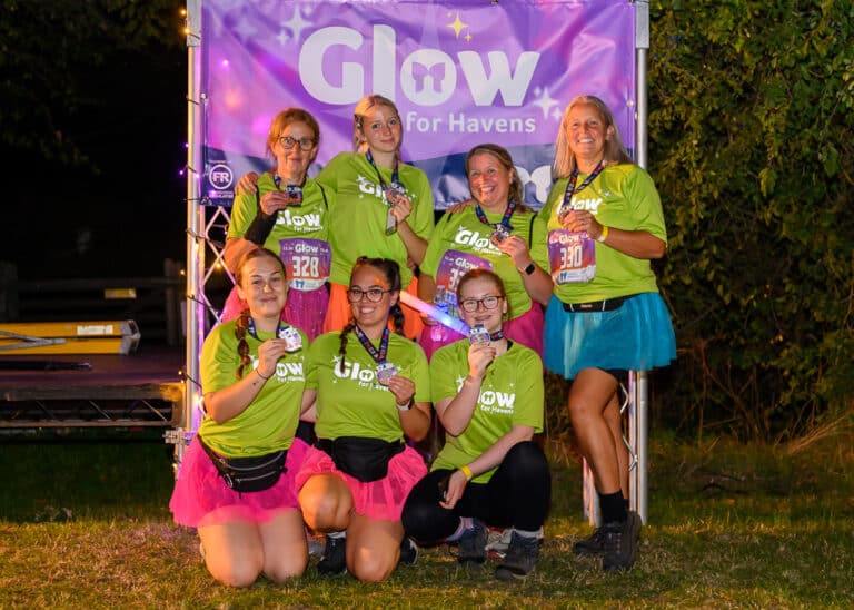 seven women smiling in bright clothing in front of a Glow for Havens banner