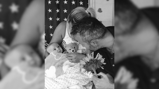 Black and white photo of baby in a blanket with his teddy and parents around him