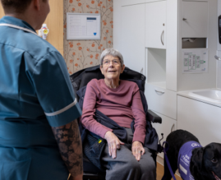 Lady sat in wheelchair with black assistance dog next to her and HCA stood talking to her with her back to the camera