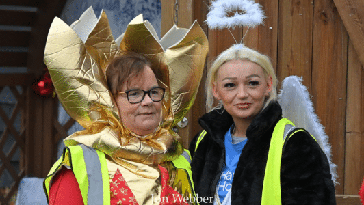 two adults dressed in festive outfits one with an angel halo and one as christmas cracker