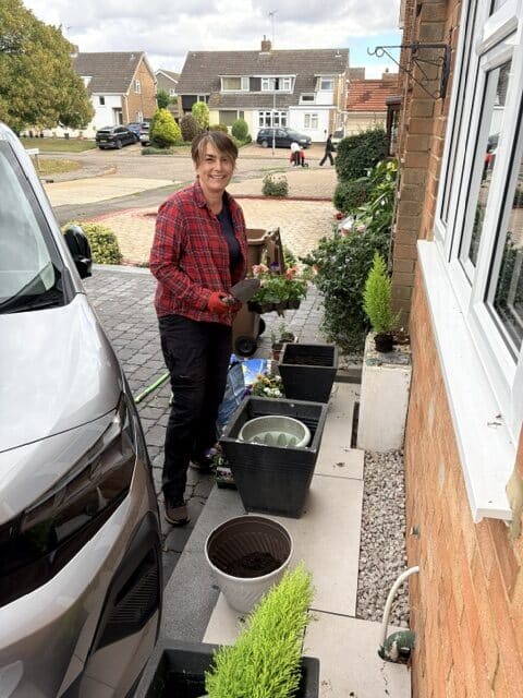 A lady smiling on someones driveway doing gardening.